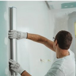 Ion popescu - Pracownik w białych rękawicach wygładza ścianę długą szpachlą. W tle okno z żółtą taśmą malarską i surowy sufit z płyt karton-gips.