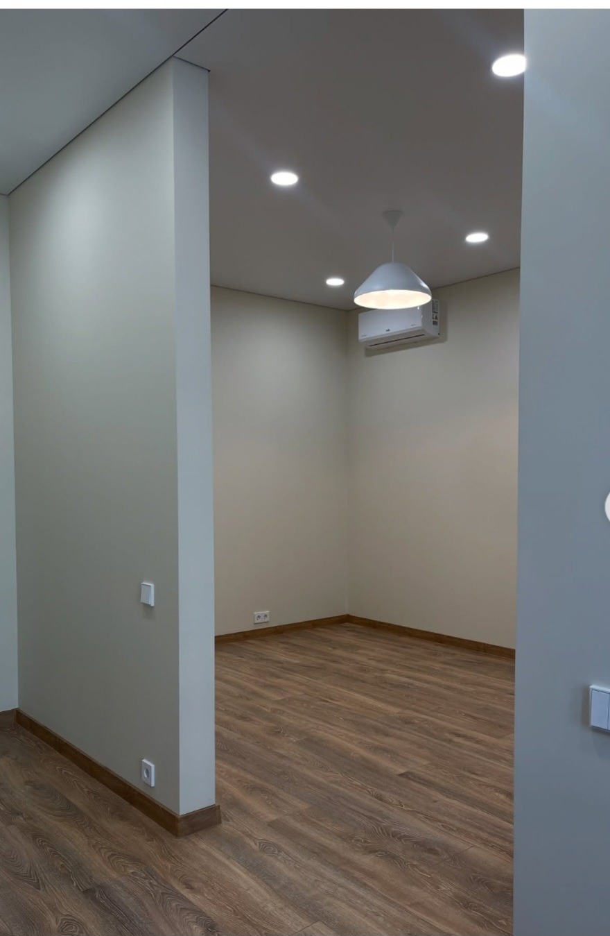 Jasne wnętrze po remoncie: drewnopodobna podłoga, kremowe ściany, biała lampa wisząca i klimatyzator. Widoczne gniazdka i włączniki.