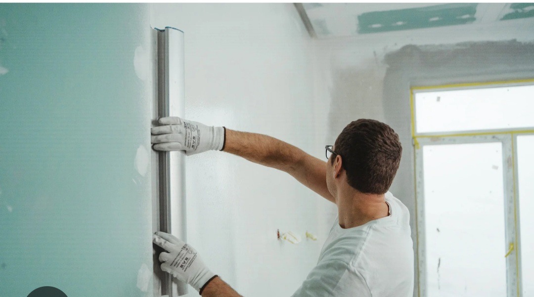 Pracownik w białych rękawicach wygładza ścianę długą szpachlą. W tle okno z żółtą taśmą malarską i surowy sufit z płyt karton-gips.