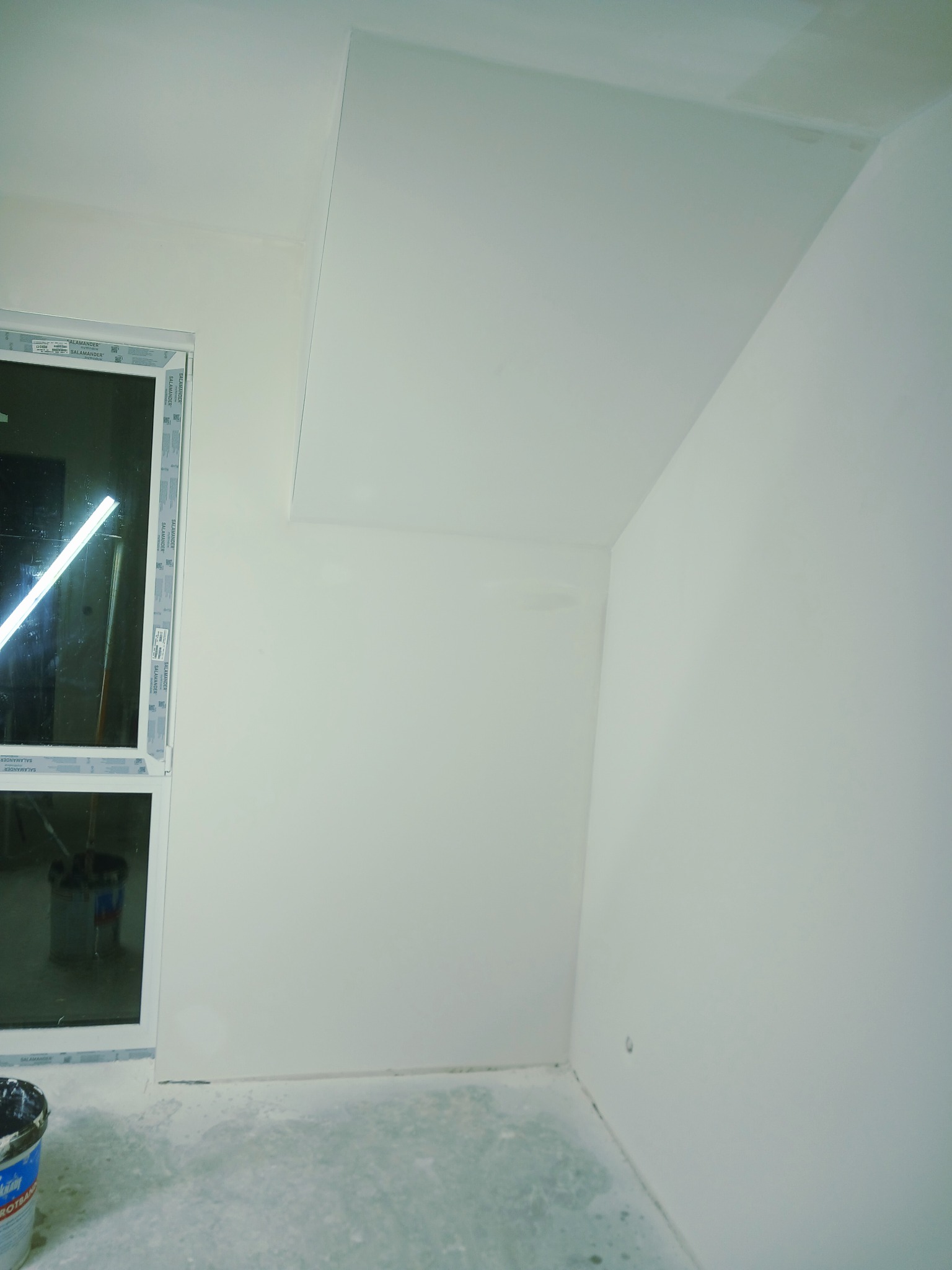 Jasne wnętrze po gładziowaniu ścian i sufitu, widoczne okno i wiadro z farbą. Minimalistyczne wykończenie, gotowe do dalszych prac.
