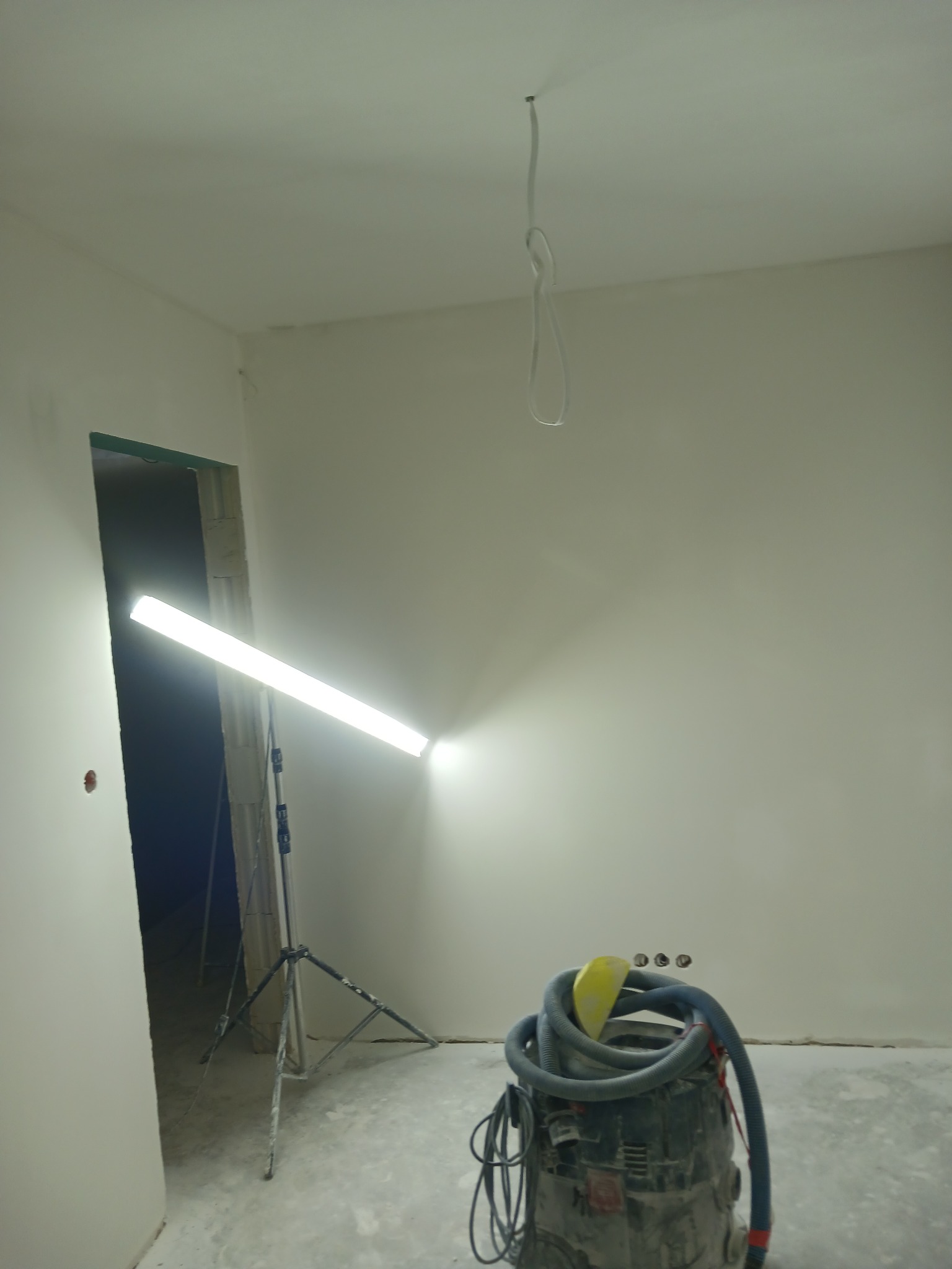 Wnętrze w trakcie prac wykończeniowych: lampa halogenowa oświetla świeżo wyszpachlowaną ścianę, obok stoi odkurzacz przemysłowy. Widoczne gniazdka elektryczne.