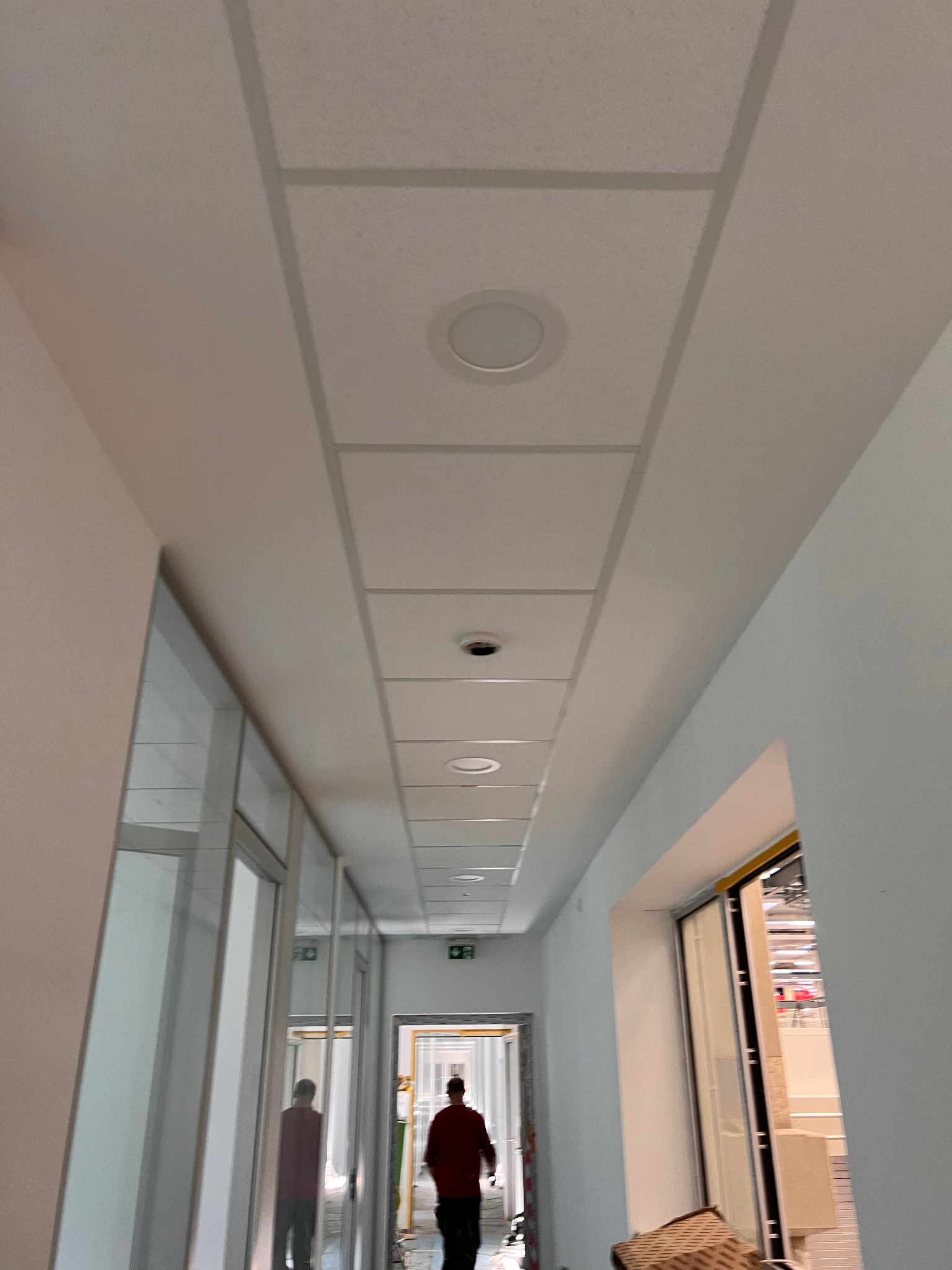 Korytarz biurowy z podwieszanym sufitem kasetonowym i oświetleniem punktowym. Widoczne szklane ścianki działowe i osoba w oddali.