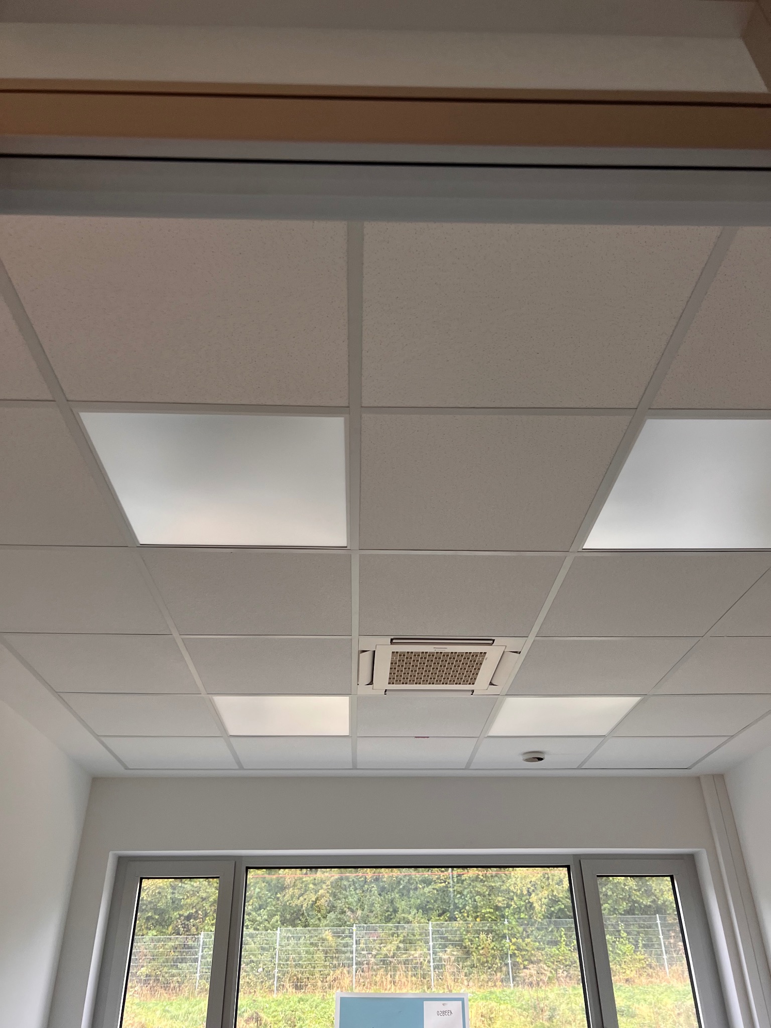 Kasetonowy sufit podwieszany z wbudowanym oświetleniem i klimatyzacją w jasnym pomieszczeniu biurowym z widokiem na zieleń za oknem.