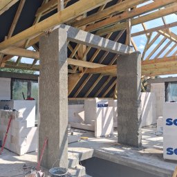 M & M SYSTEM - Wnętrze domu w trakcie budowy: widoczne ściany z bloczków, drewniana konstrukcja dachu, betonowe filary. Na podłodze narzędzia i materiały budowlane.