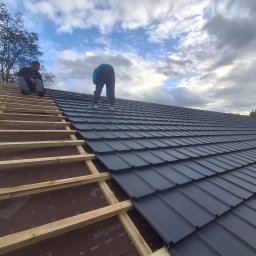 M & M SYSTEM - Montaż blachodachówki na drewnianej konstrukcji dachu. Dwóch mężczyzn pracuje na dachu, jeden siedzi, drugi montuje. Widok na pochmurne niebo.