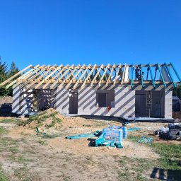 M & M SYSTEM - Budowa domu z białych bloczków w Nieporęcie, widoczne drewniane elementy konstrukcji dachu i pracownicy na górze. Na pierwszym planie materiały budowlane.