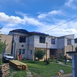 M & M SYSTEM - Niedokończony dom z białego pustaka, zabezpieczony niebieską plandeką na dachu. Na pierwszym planie uszkodzony samochód i stos drewna. W tle błękitne niebo z chmurami.