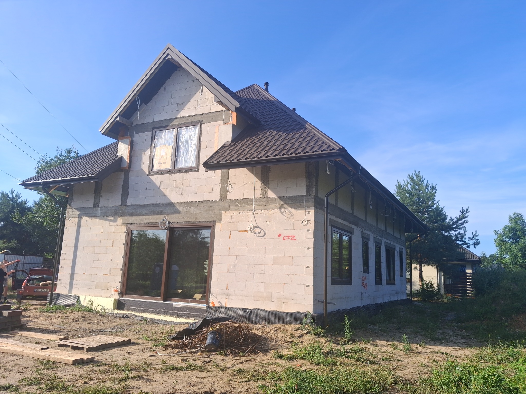 Dwukondygnacyjny dom w trakcie budowy z białych bloczków, z brązowym dachem i oknami. Widoczne instalacje elektryczne i materiały budowlane na posesji. Słoneczny dzień.