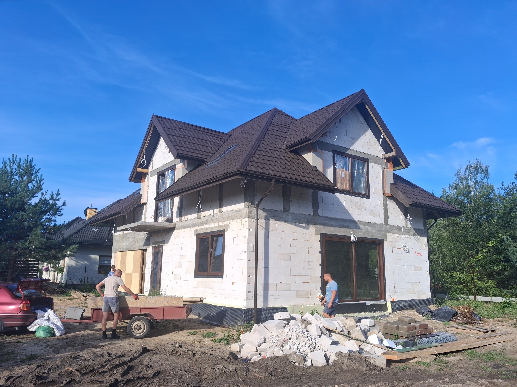 Dwukondygnacyjny dom w trakcie budowy z białego bloczka, z brązowym dachem. Widoczni dwaj mężczyźni i przyczepka. Na pierwszym planie gruz i materiały budowlane.