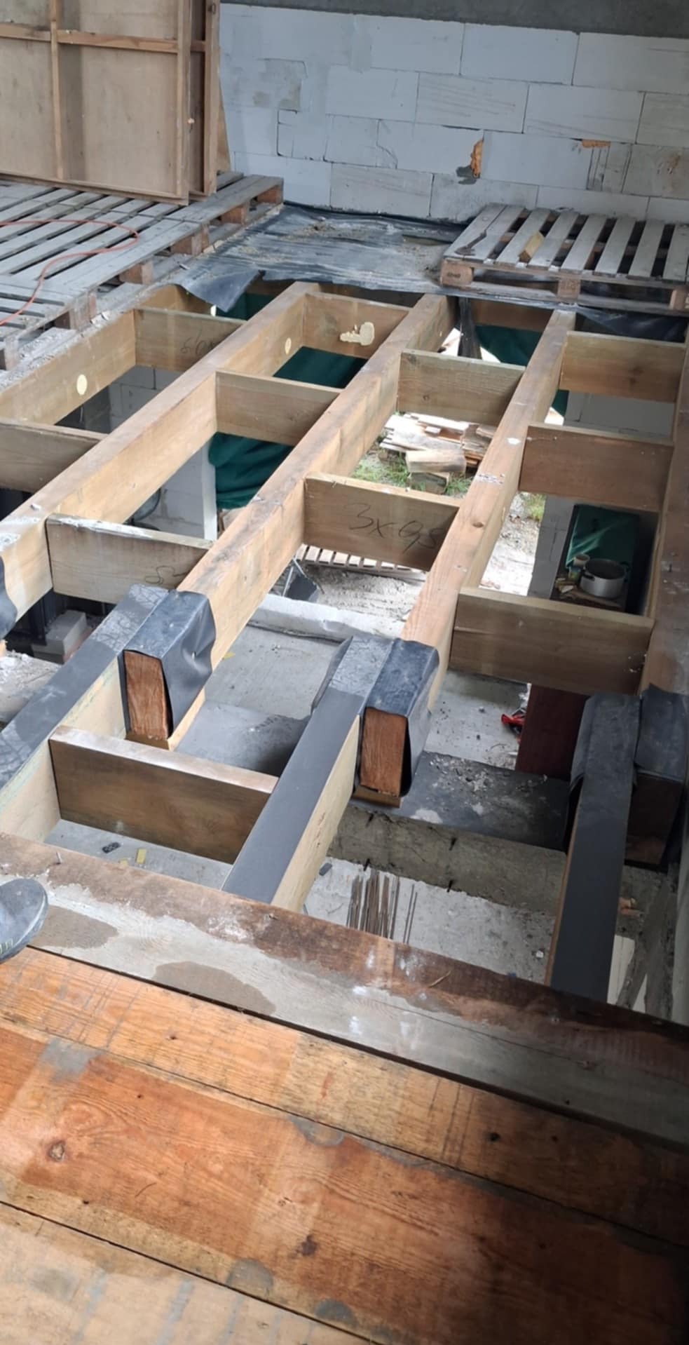 Drewniana konstrukcja pod podłogę z izolacją, widoczne bloczki betonowe w tle. Etap budowy z bliska, z naciskiem na detale łączeń i materiałów.