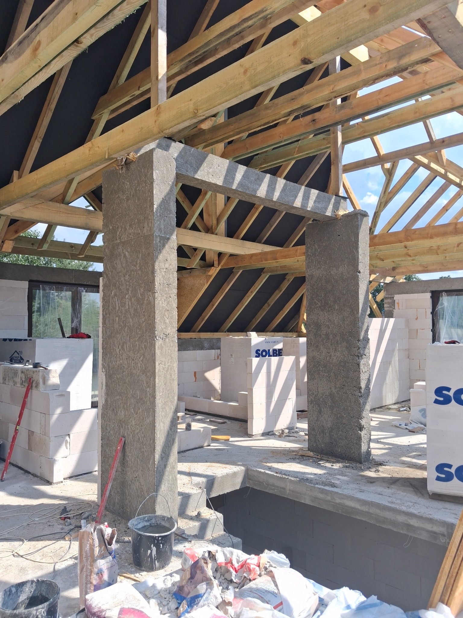 Wnętrze domu w trakcie budowy: widoczne ściany z bloczków, drewniana konstrukcja dachu, betonowe filary. Na podłodze narzędzia i materiały budowlane.