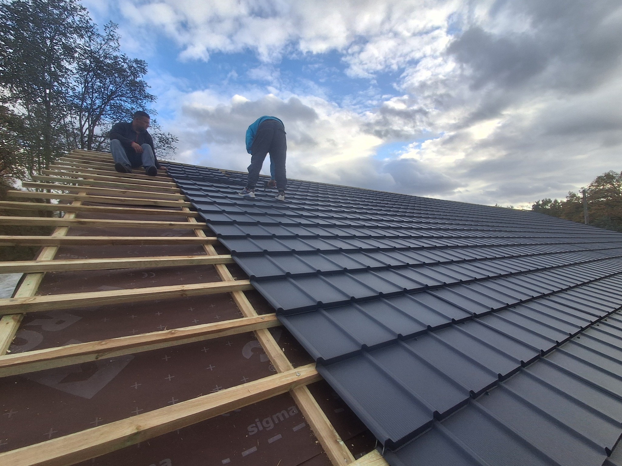 Montaż blachodachówki na drewnianej konstrukcji dachu. Dwóch mężczyzn pracuje na dachu, jeden siedzi, drugi montuje. Widok na pochmurne niebo.