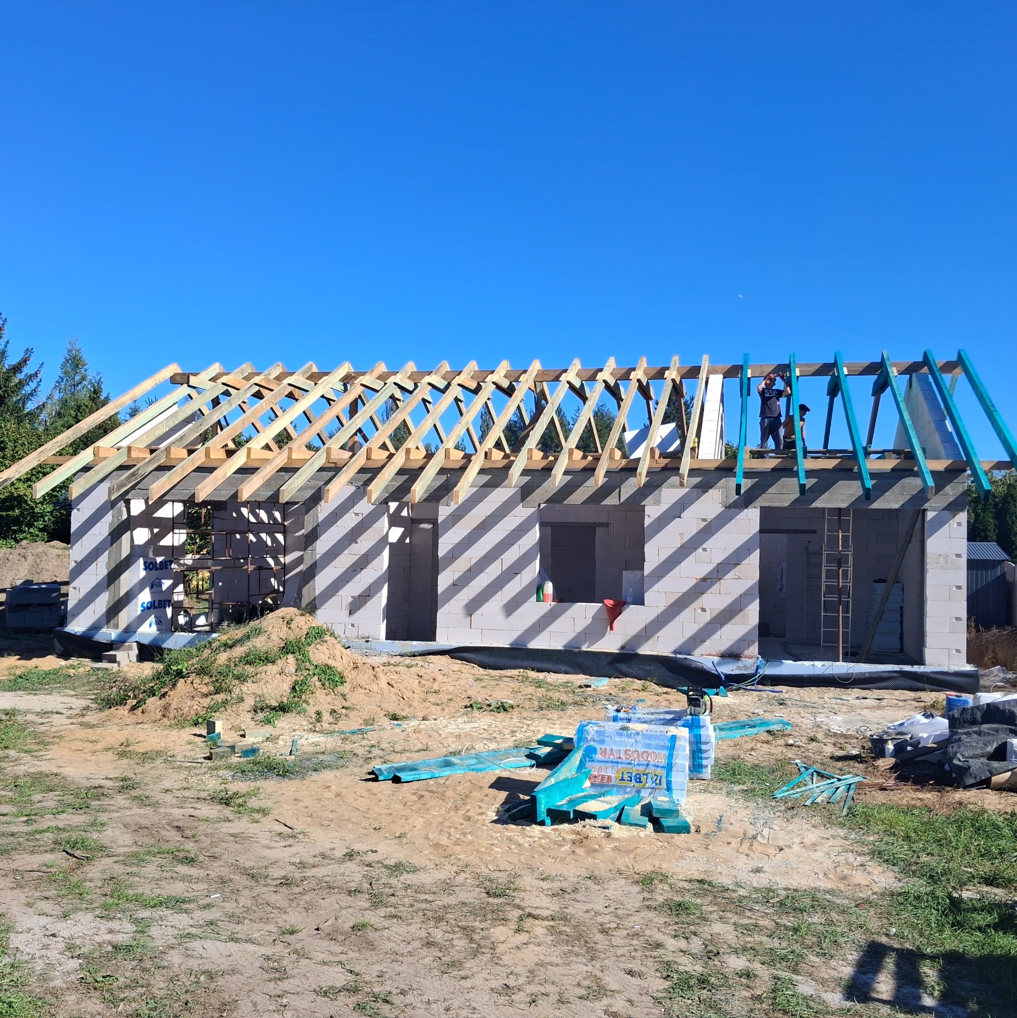 Budowa domu z białych bloczków w Nieporęcie, widoczne drewniane elementy konstrukcji dachu i pracownicy na górze. Na pierwszym planie materiały budowlane.