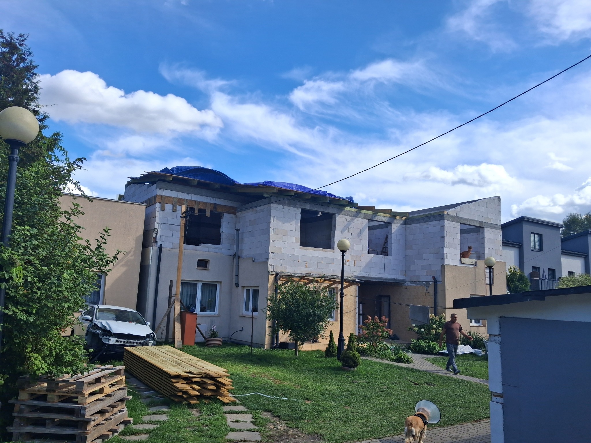 Niedokończony dom z białego pustaka, zabezpieczony niebieską plandeką na dachu. Na pierwszym planie uszkodzony samochód i stos drewna. W tle błękitne niebo z chmurami.