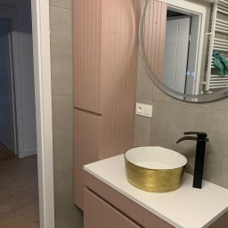 Danch Danylo Muzyka - Nowoczesna łazienka z pudrowo-różową zabudową meblową, złotą umywalką i czarną armaturą. Na ścianie szare płytki i okrągłe lustro z podświetleniem LED.