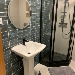 Danch Danylo Muzyka - Nowoczesna łazienka z kabiną prysznicową w czarnej ramie, umywalką na postumencie i okrągłym lustrem z podświetleniem. W tle płytki w odcieniach szarości. W rogu wiertarka.