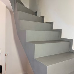 Danch Danylo Muzyka - Pomalowane na szaro schody wewnątrz budynku, widok z dołu. Ściany białe i jasnoróżowe, podłoga drewnopodobna. Widoczne gniazdko elektryczne i fragment odkurzacza.