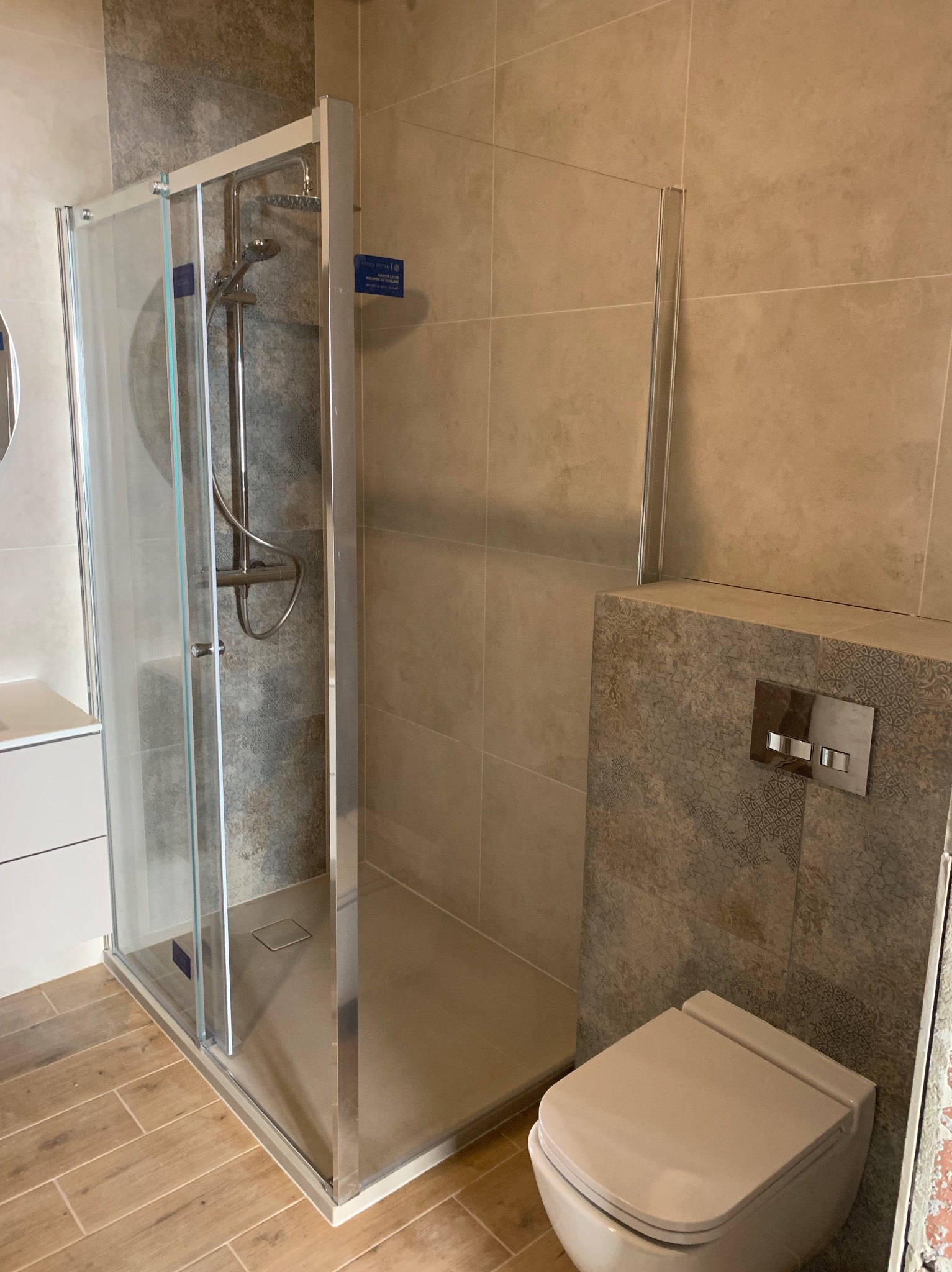 Nowoczesna kabina prysznicowa z chromowanymi elementami i szklanymi drzwiami, beżowe płytki na ścianach, drewnopodobna podłoga, wisząca toaleta z przyciskiem spłukującym.