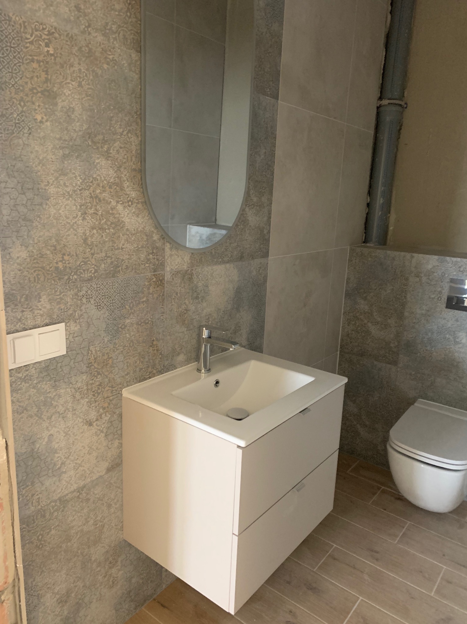 Wykończona łazienka z umywalką na szafce, lustrem i toaletą. Płytki ścienne w odcieniach szarości z wzorem, drewnopodobne płytki podłogowe. Widoczna instalacja kanalizacyjna.