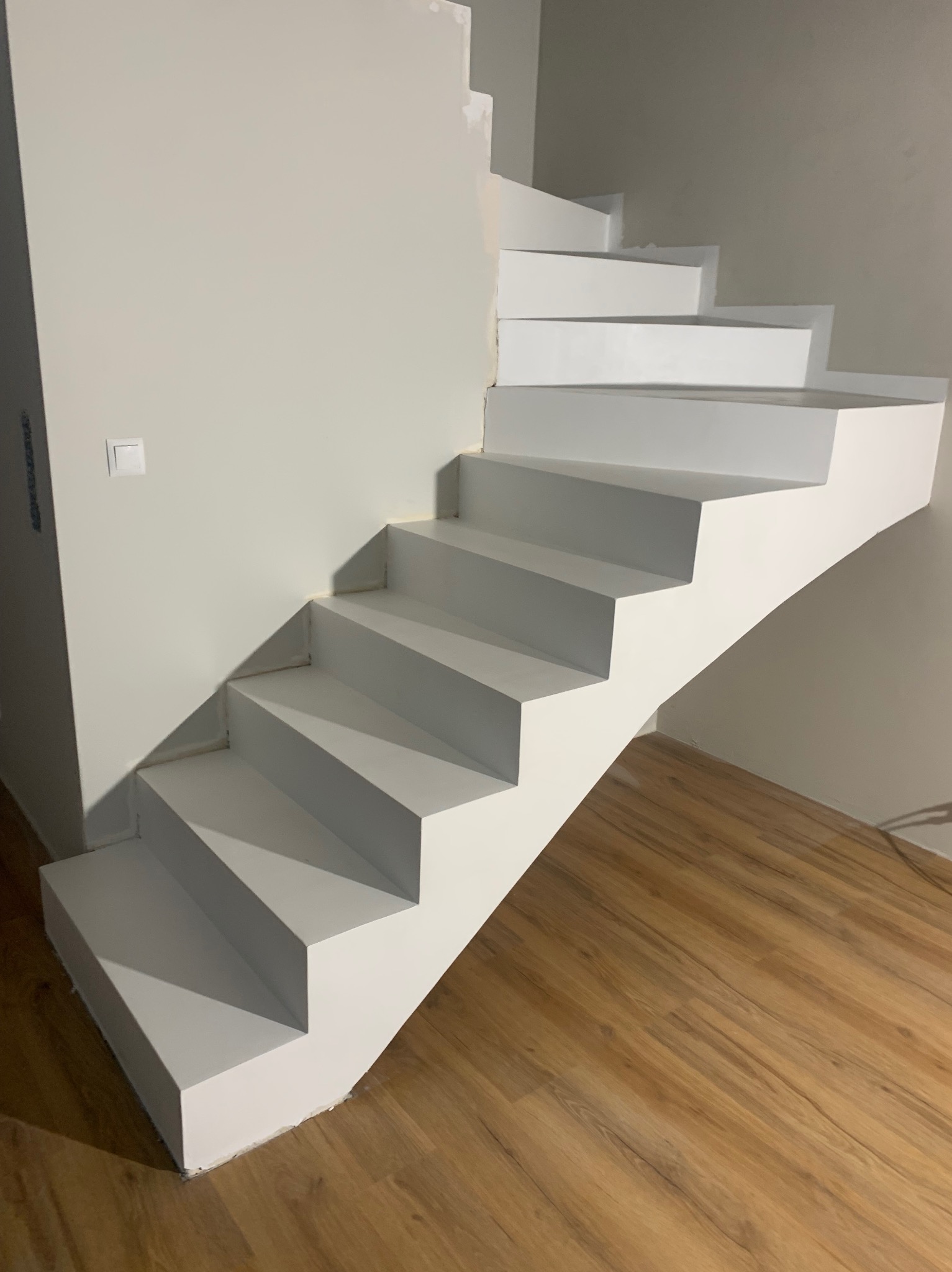 Białe, nowoczesne schody spiralne z drewnianą podłogą i szarą ścianą. Minimalistyczny design, widoczne detale wykończenia. Oświetlenie naturalne.