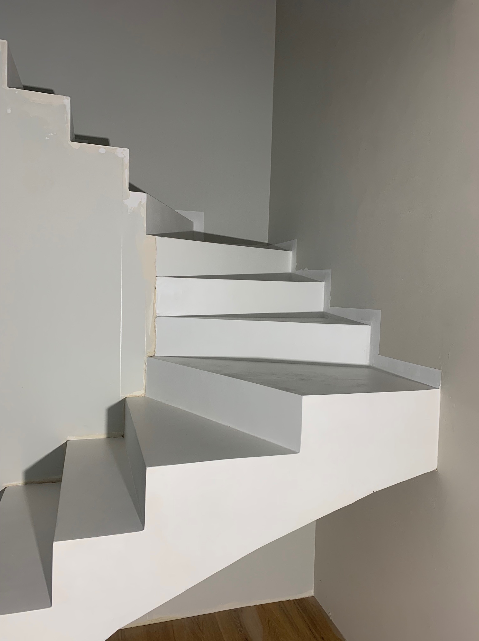 Białe, betonowe schody spiralne w trakcie malowania, widoczne niedociągnięcia i szpachlowanie na ścianie. Minimalistyczny design, surowe wykończenie.