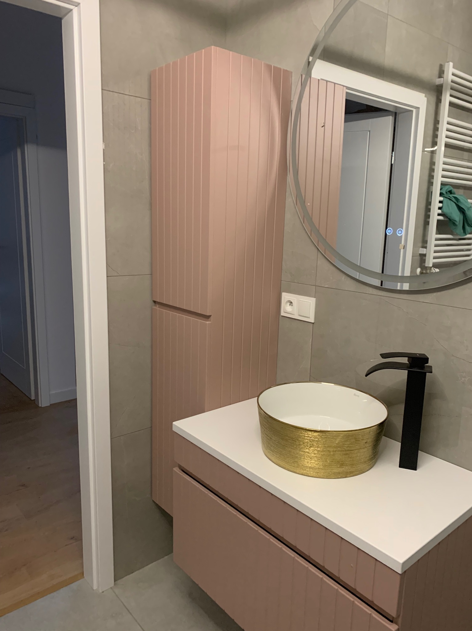 Nowoczesna łazienka z pudrowo-różową zabudową meblową, złotą umywalką i czarną armaturą. Na ścianie szare płytki i okrągłe lustro z podświetleniem LED.