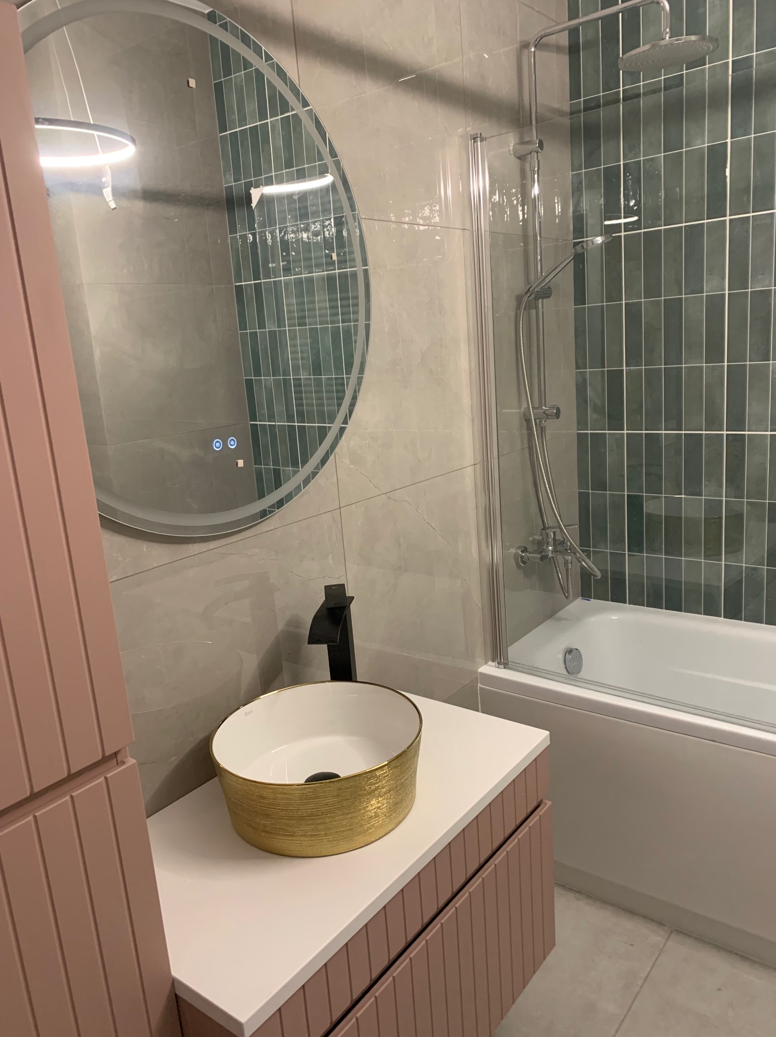 Nowoczesna łazienka po remoncie: złota umywalka nablatowa, okrągłe lustro z podświetleniem LED, kabina prysznicowa nad wanną i płytki w odcieniach szarości i zieleni.