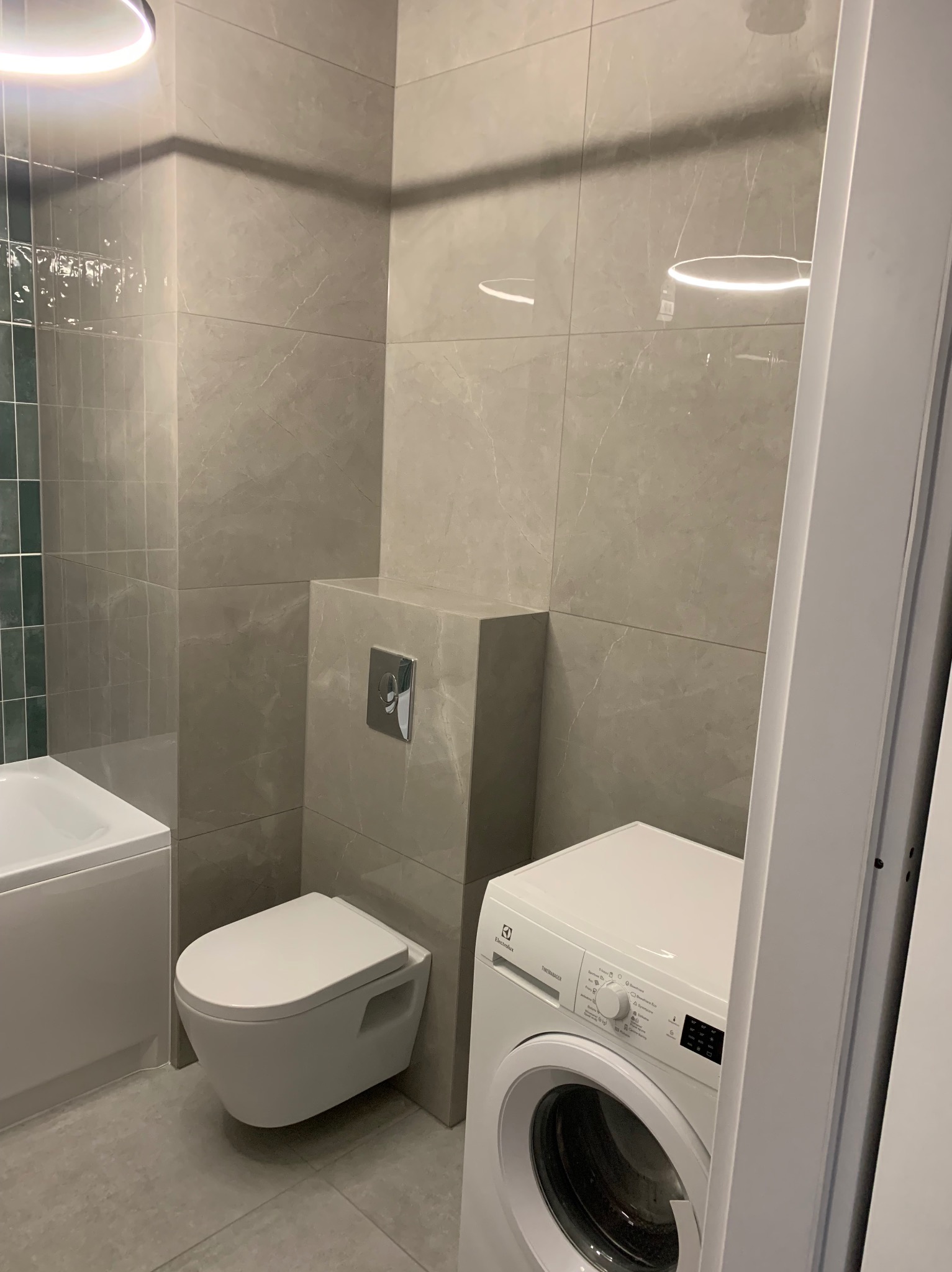 Wykończona łazienka w szarościach: podwieszana toaleta, pralka Electrolux, płytki imitujące marmur, oświetlenie LED. Minimalistyczny design, nowoczesne rozwiązania.