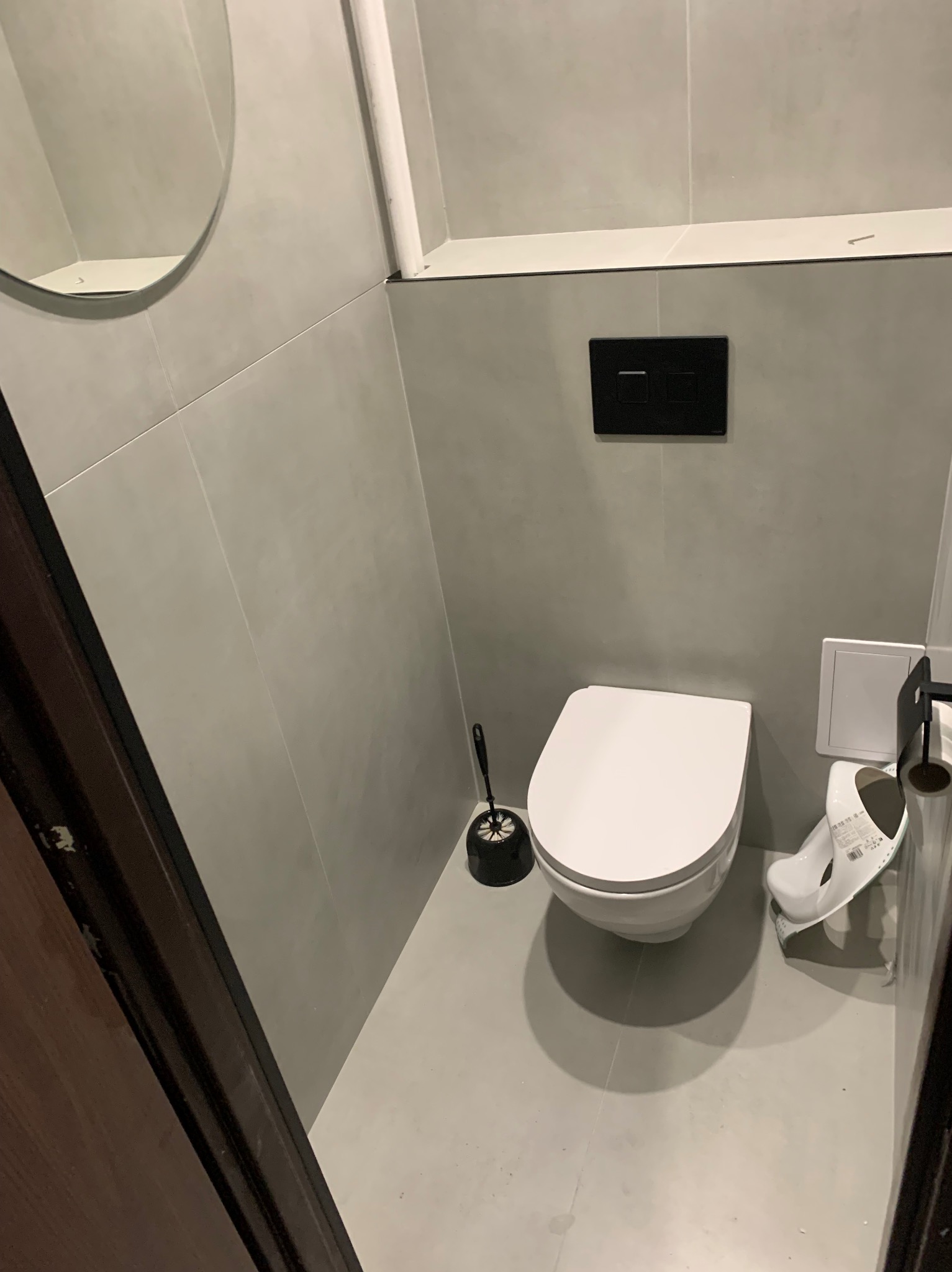 Minimalistyczna toaleta w szarościach z okrągłym lustrem i czarnym przyciskiem spłukiwania, widok z perspektywy drzwi, po remoncie.