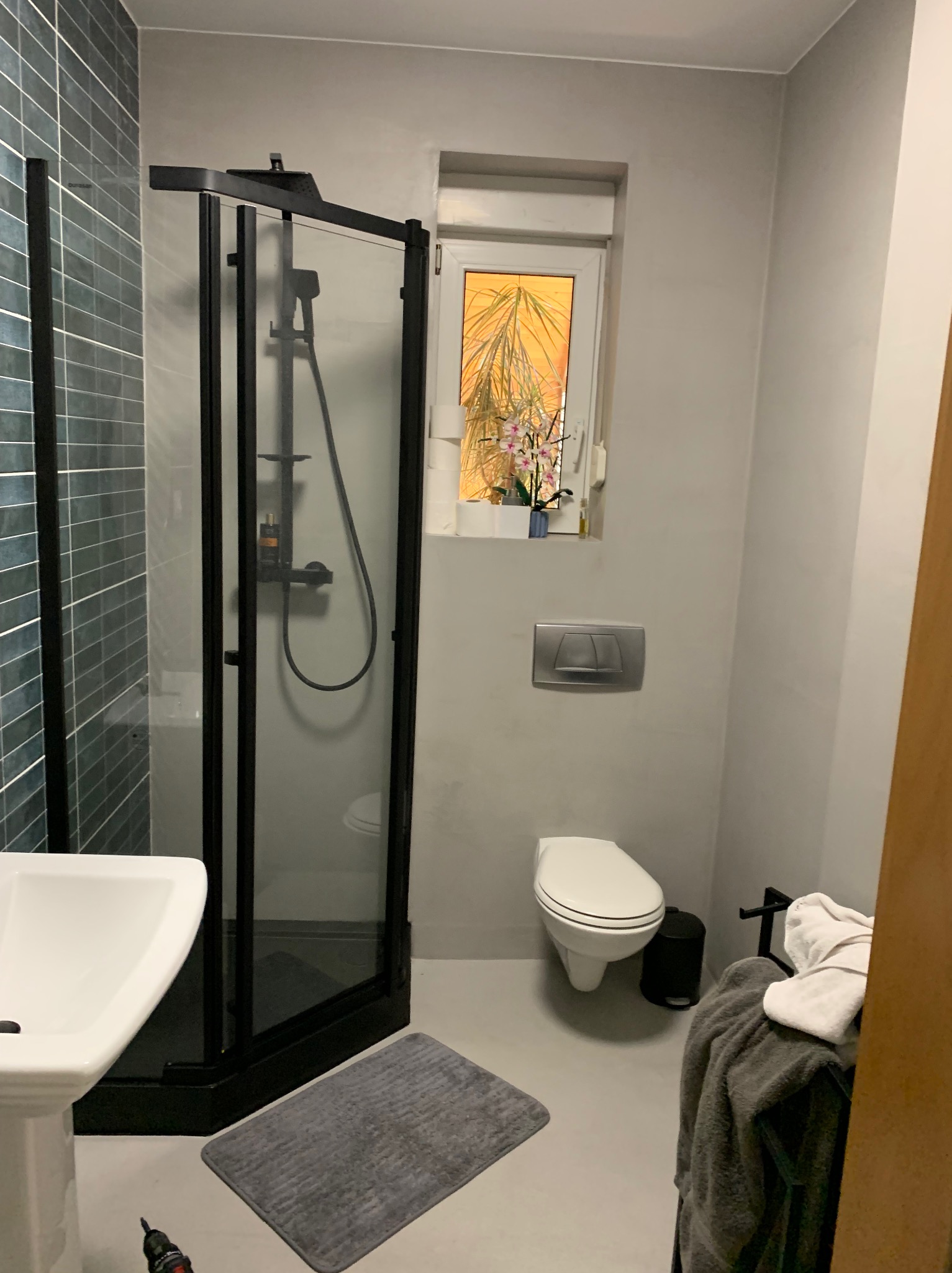 Nowoczesna łazienka po remoncie w Poznaniu. Kabina prysznicowa z czarnymi ramami, umywalka, toaleta i szare płytki. Minimalistyczny design z roślinami w oknie.