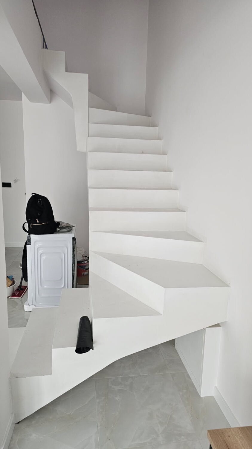 Białe, minimalistyczne schody w nowoczesnym wnętrzu. Widoczna pralka i plecak w tle. Jasne płytki podłogowe i białe ściany tworzą spójną całość.