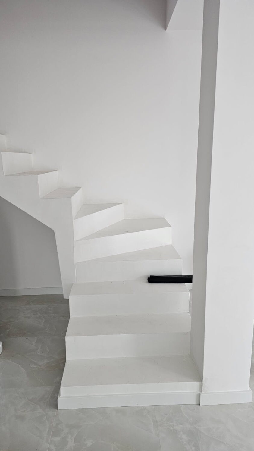 Minimalistyczne, białe schody w nowoczesnym wnętrzu. Widoczne marmurowe płytki podłogowe i czarny element na schodach. Ściany i kolumna również w bieli.