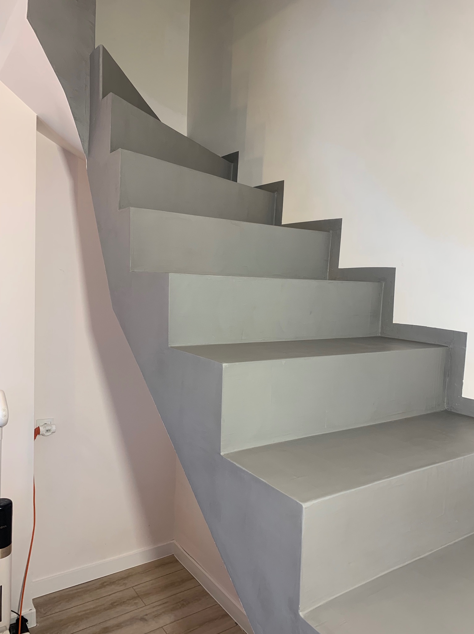 Pomalowane na szaro schody wewnątrz budynku, widok z dołu. Ściany białe i jasnoróżowe, podłoga drewnopodobna. Widoczne gniazdko elektryczne i fragment odkurzacza.