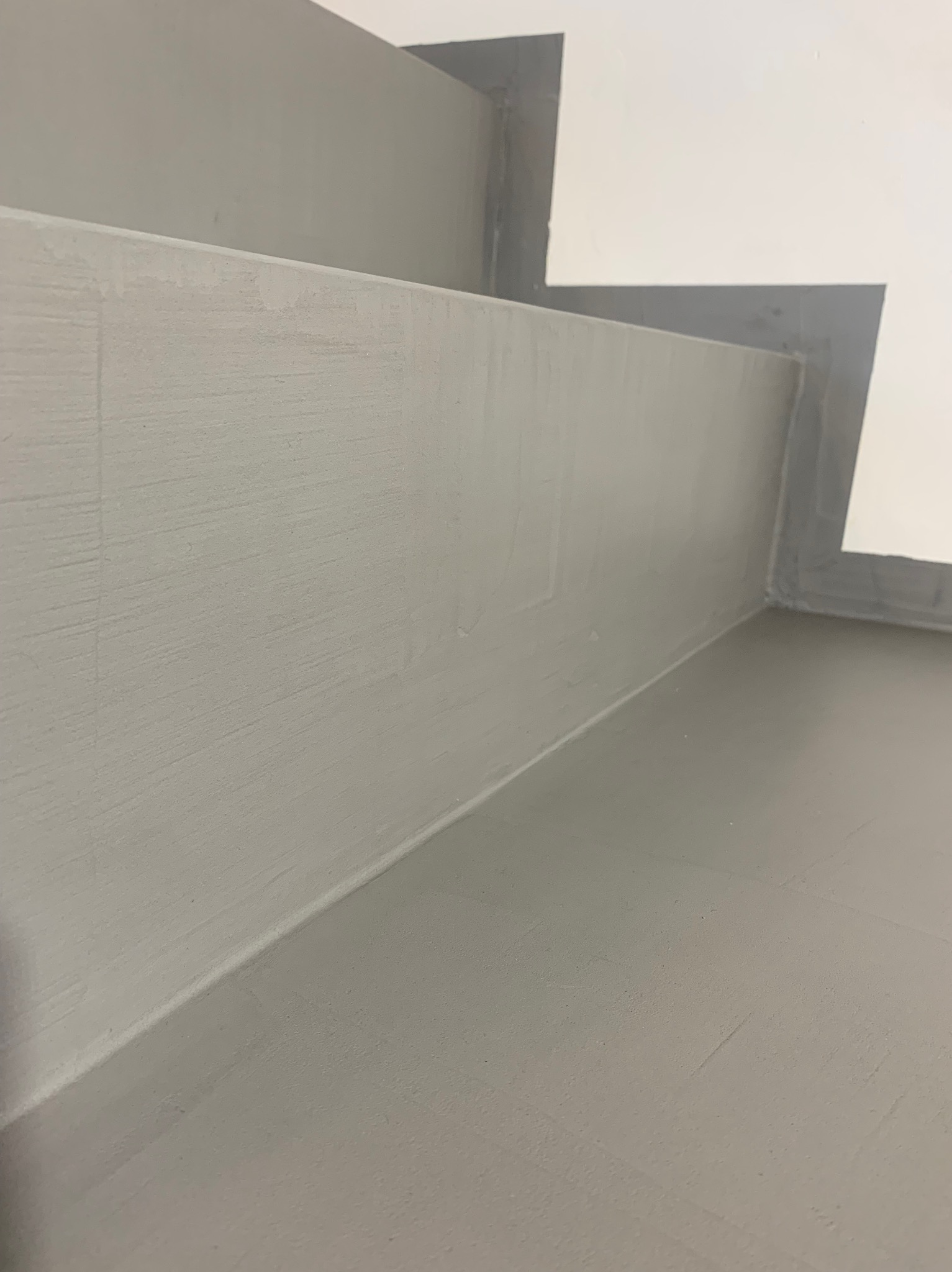 Szare, świeżo pomalowane ściany i podłoga w rogu pomieszczenia, widoczne ślady pędzla, biały fragment ściany w górnym rogu. Minimalistyczne wykończenie.