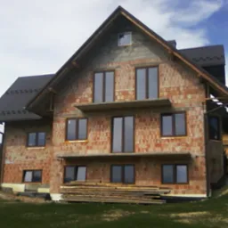 Surowy, dwupiętrowy dom z czerwonej cegły w trakcie budowy, z zamontowanymi oknami i częściowo pokrytym dachem, na tle zielonego wzgórza i pochmurnego nieba.