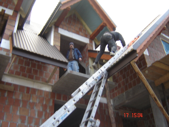 Budowa domu z cegły: pracownik na drabinie montuje konstrukcję dachu, drugi pracownik obserwuje z balkonu, widoczne rusztowania i nieukończone elementy konstrukcyjne.