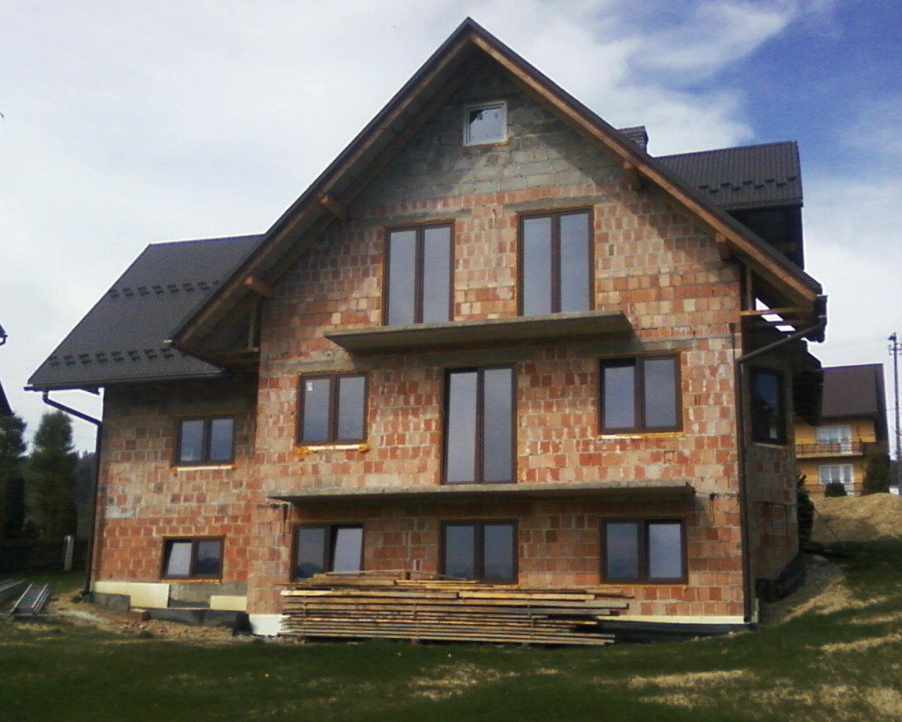 Surowy, dwupiętrowy dom z czerwonej cegły w trakcie budowy, z zamontowanymi oknami i częściowo pokrytym dachem, na tle zielonego wzgórza i pochmurnego nieba.