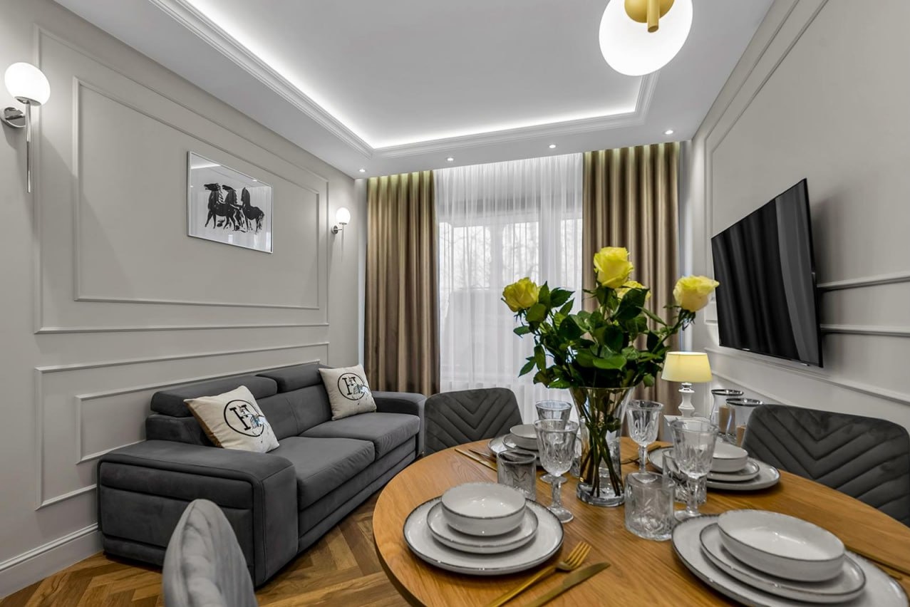 Eleganckie wnętrze: szary narożnik z poduszkami, okrągły stół z zastawą, żółte róże w wazonie, telewizor na ścianie i dekoracyjne listwy ścienne.