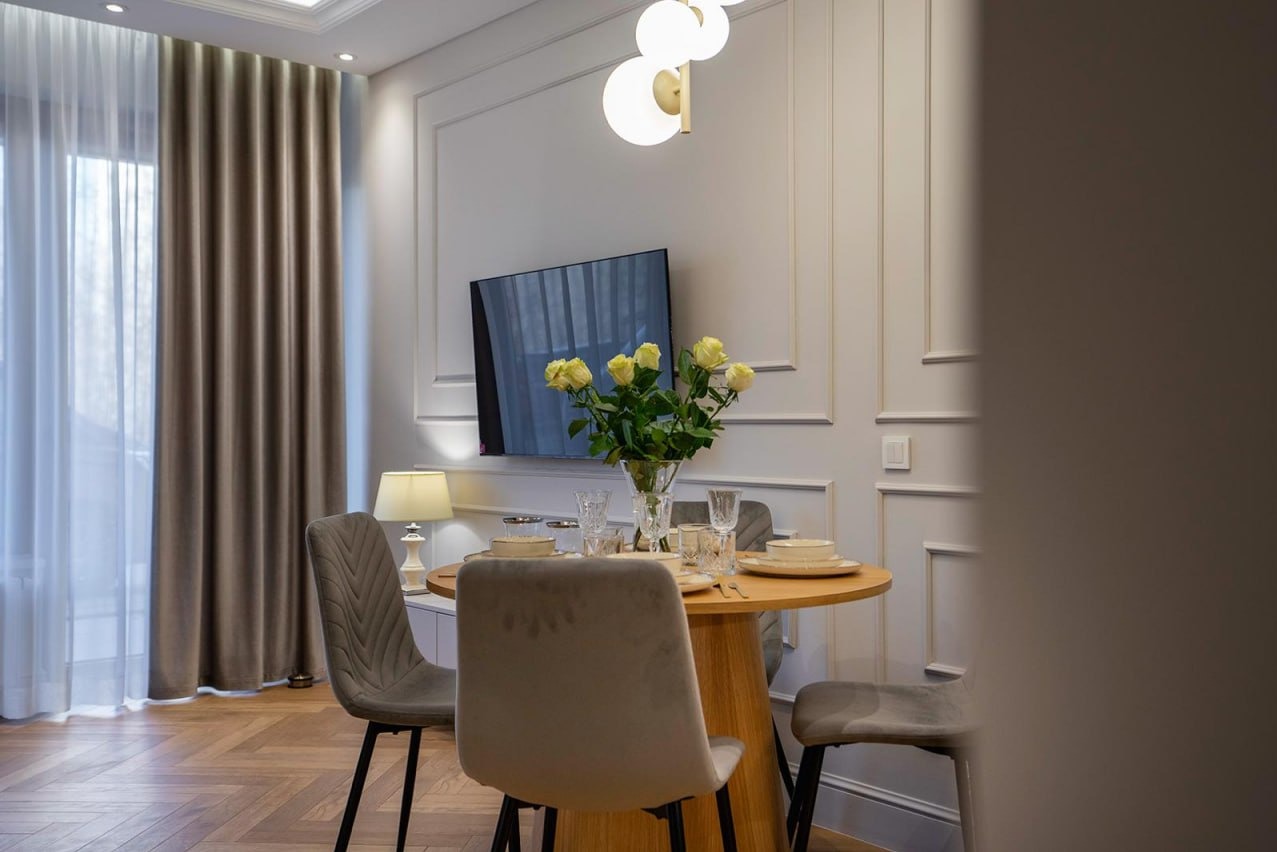 Elegancka jadalnia: okrągły stół z bukietem żółtych róż, szare krzesła, telewizor na ścianie z dekoracyjnymi panelami, parkiet w jodełkę i zasłony w tle.