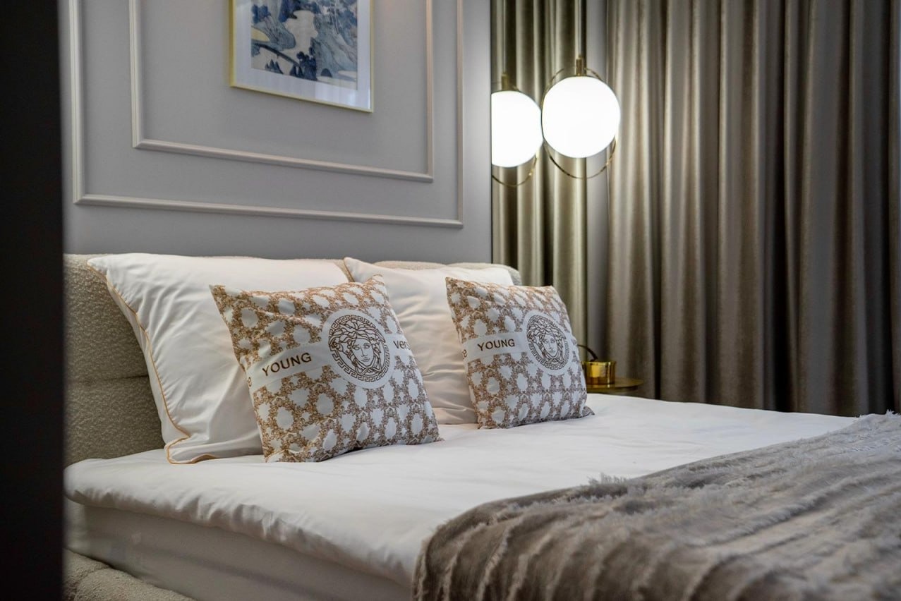 Elegancka sypialnia z łóżkiem, dekoracyjnymi poduszkami Versace Young, lampą wiszącą i zasłonami w odcieniach szarości. Na ścianie obraz w ramie.