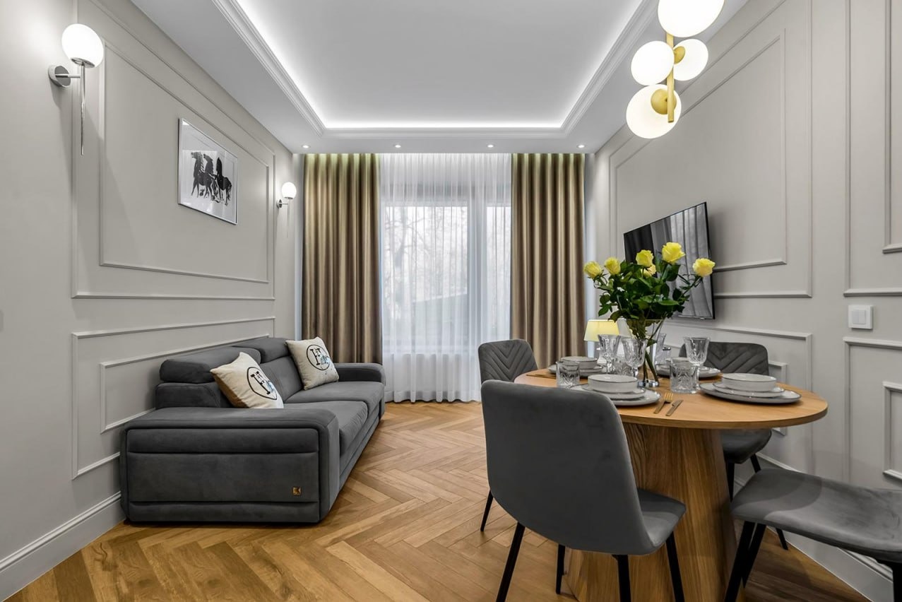 Stylowe wnętrze salonu z szarą sofą, okrągłym stołem i parkietem w jodełkę. Ściany zdobione sztukaterią, nowoczesne oświetlenie i żółte róże w wazonie.