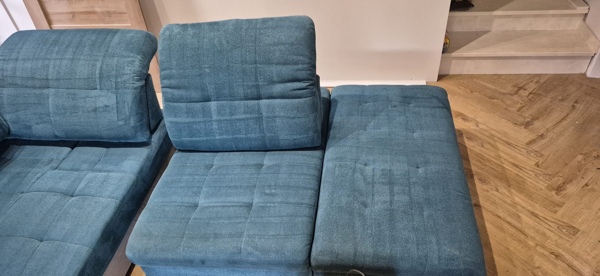 Turkusowa, modułowa kanapa z tkaniny w zbliżeniu, widoczne pikowania i faktura materiału. Parkiet w jodełkę i fragment schodów w tle. Stonowana kolorystyka.