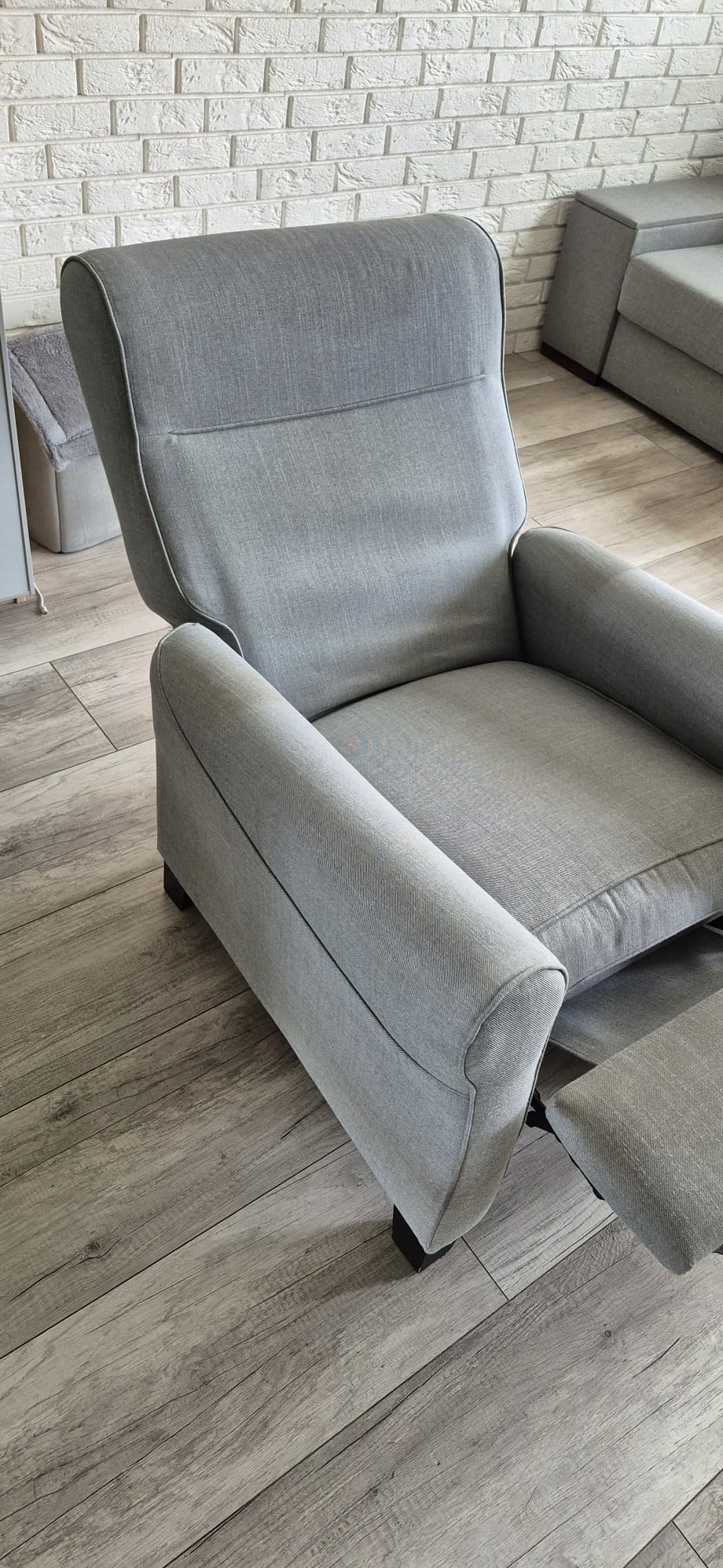 Szary, rozkładany fotel tapicerowany z podnóżkiem na tle drewnianej podłogi i białej ściany z cegły. Widoczny fragment sofy w tle.