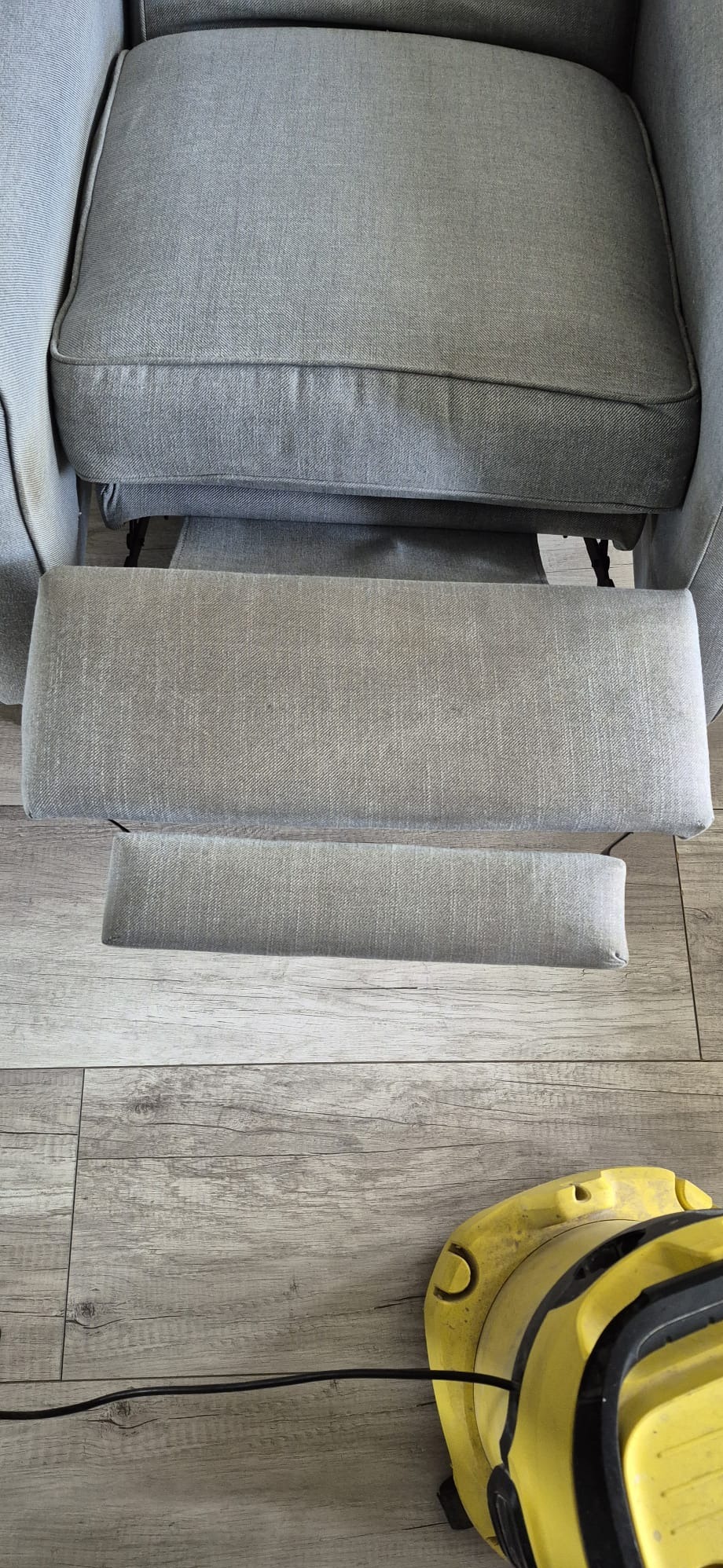 Szary fotel z wysuniętym podnóżkiem, widoczny fragment żółtego odkurzacza piorącego na drewnianej podłodze. Ujęcie z góry, skupione na tapicerce.
