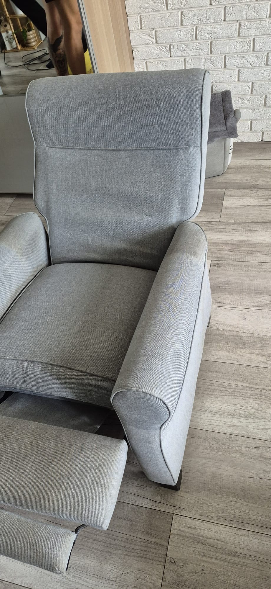 Szary, tapicerowany fotel z podnóżkiem na drewnianej podłodze. Widok z tyłu, z elementami otoczenia: fragment ściany z cegły i lustro. Czysty, jasny kadr.