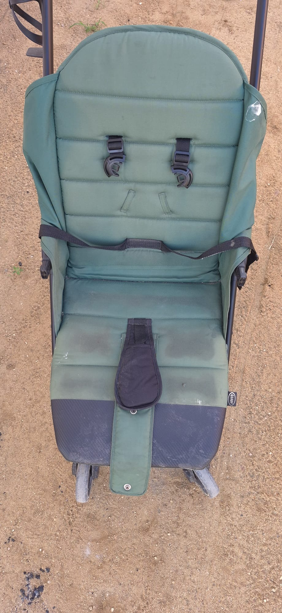 Zielony wózek dziecięcy z widocznymi plamami na tapicerce, stojący na piasku. Widoczne pasy bezpieczeństwa i elementy konstrukcyjne wózka.