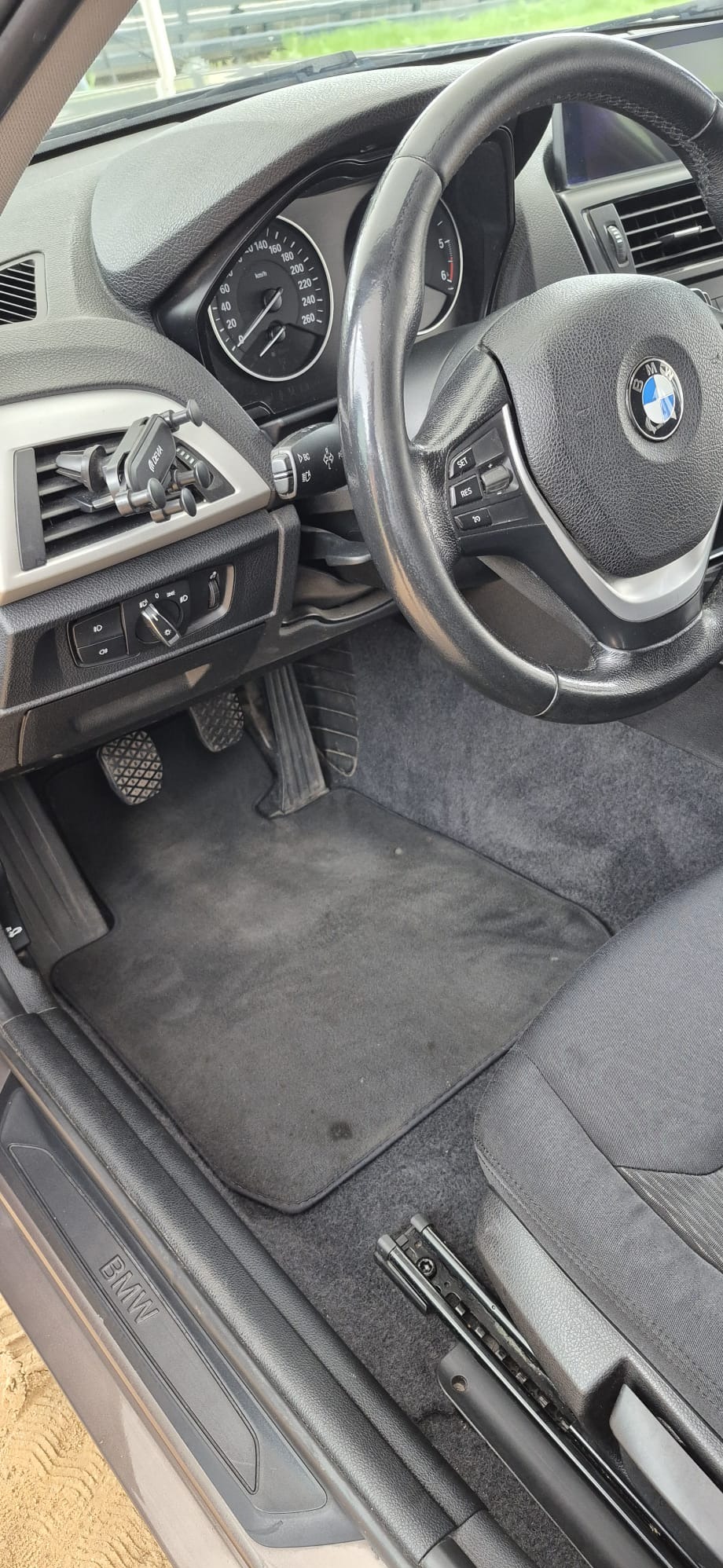Wnętrze samochodu BMW z widocznymi pedałami, dywanikiem z plamą i fragmentem fotela. Kierownica z logo BMW w górnej części kadru.