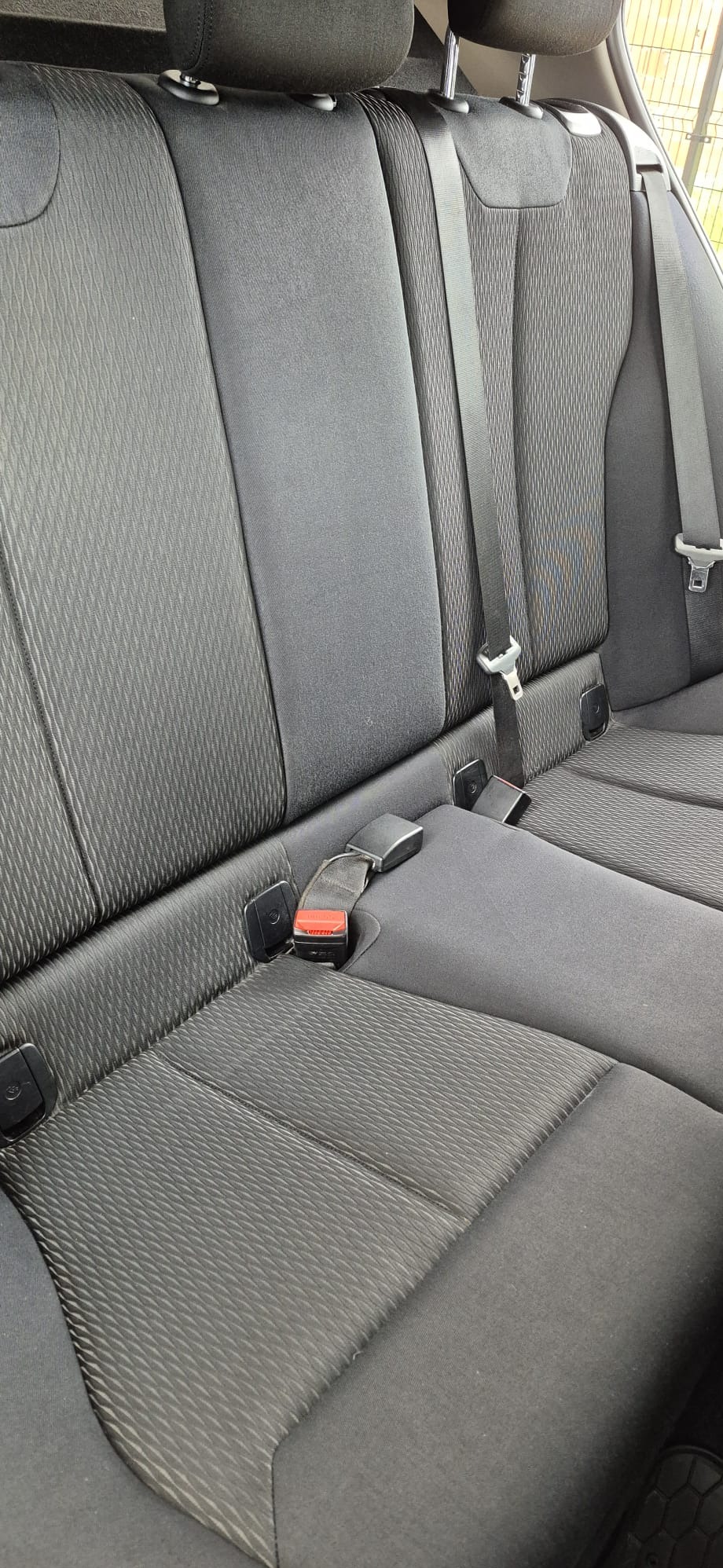 Szara, tekstylna tapicerka tylnej kanapy samochodowej z pasami bezpieczeństwa. Widoczne zagłówki i wzór na materiale. Czyste, zadbane wnętrze pojazdu.