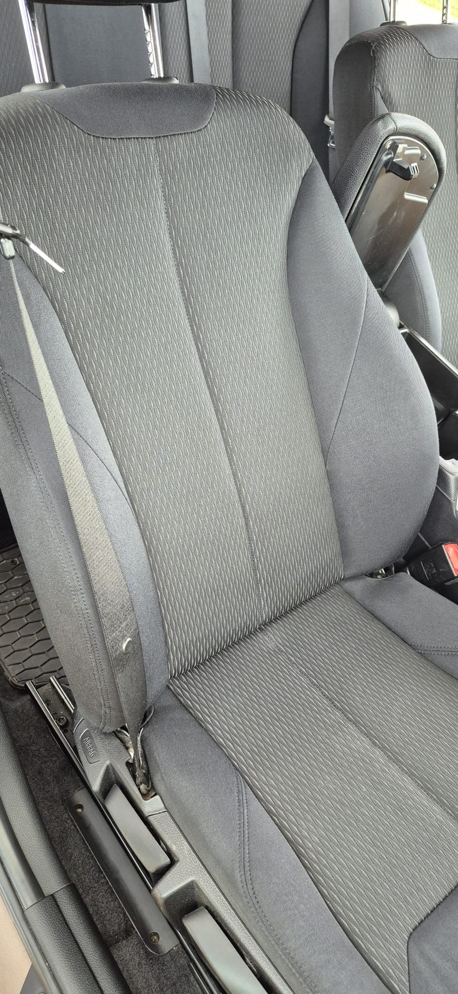 Szare, tekstylne siedzenie samochodowe z widocznym pasem bezpieczeństwa i zagłówkiem. Widoczna faktura materiału i elementy konstrukcyjne fotela.