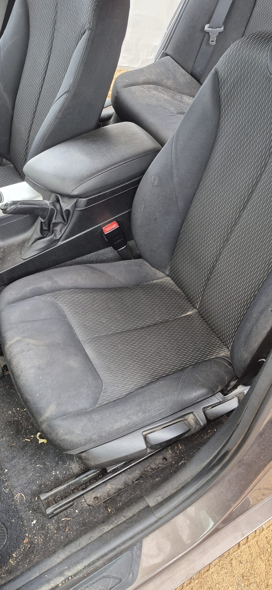 Bardzo brudne, czarne, tekstylne siedzenie samochodowe z widocznymi plamami i zabrudzeniami na tapicerce. Widoczny fragment podłogi z zanieczyszczeniami.
